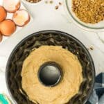 Bundt cake pan filled with batter, surrounded by cracked eggs, chopped nuts, brown sugar, a spatula, and a checkered towel on a marble countertop.