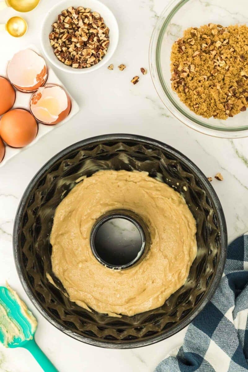 Bundt cake pan filled with batter, surrounded by cracked eggs, chopped nuts, brown sugar, a spatula, and a checkered towel on a marble countertop.
