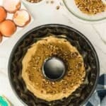 Unbaked bundt cake batter in a pan, topped with brown sugar and chopped nuts, surrounded by eggshells, a bowl of nuts, and a spatula on a marble countertop.