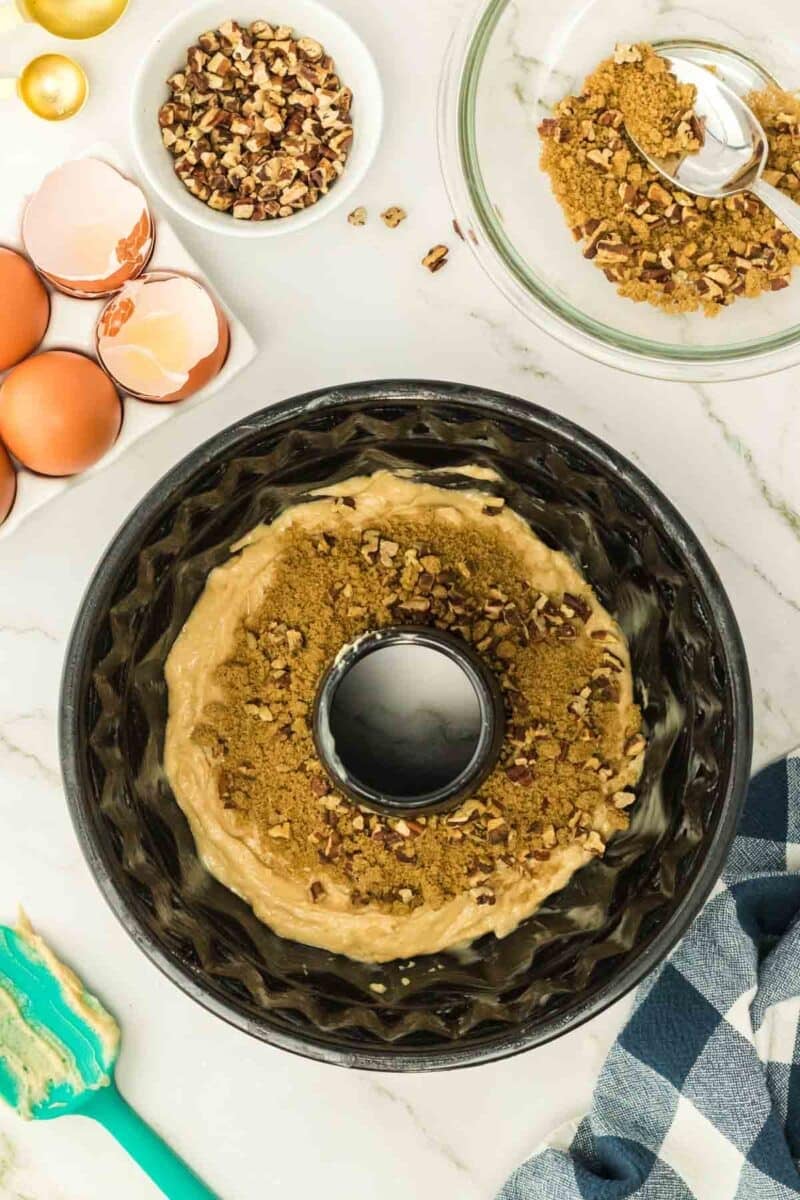 Unbaked bundt cake batter in a pan, topped with brown sugar and chopped nuts, surrounded by eggshells, a bowl of nuts, and a spatula on a marble countertop.
