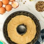 Bundt cake batter in a pan surrounded by eggs, butter cubes, chopped nuts, and a checkered towel on a marble countertop.