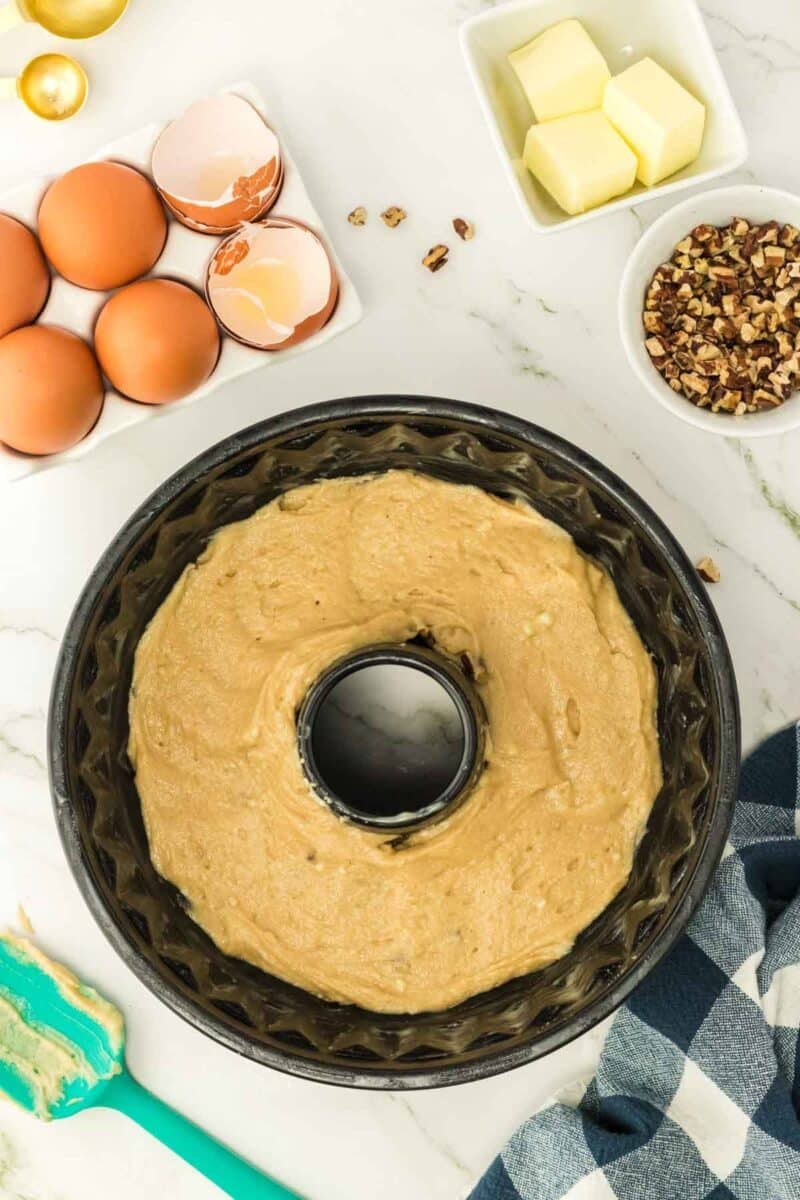 Bundt cake batter in a pan surrounded by eggs, butter cubes, chopped nuts, and a checkered towel on a marble countertop.