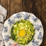 A hand pours yellow salad dressing from a spoon over chopped lettuce in a white bowl with blue floral designs, on a wooden table.