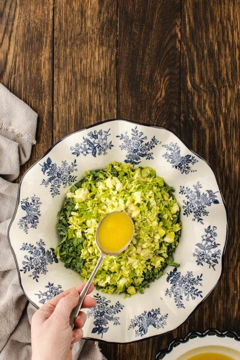 A hand pours yellow salad dressing from a spoon over chopped lettuce in a white bowl with blue floral designs, on a wooden table.