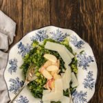 Salad with chopped greens, apple slices, and shaved cheese is served in a white bowl with blue floral patterns on a wooden table, with two serving utensils.