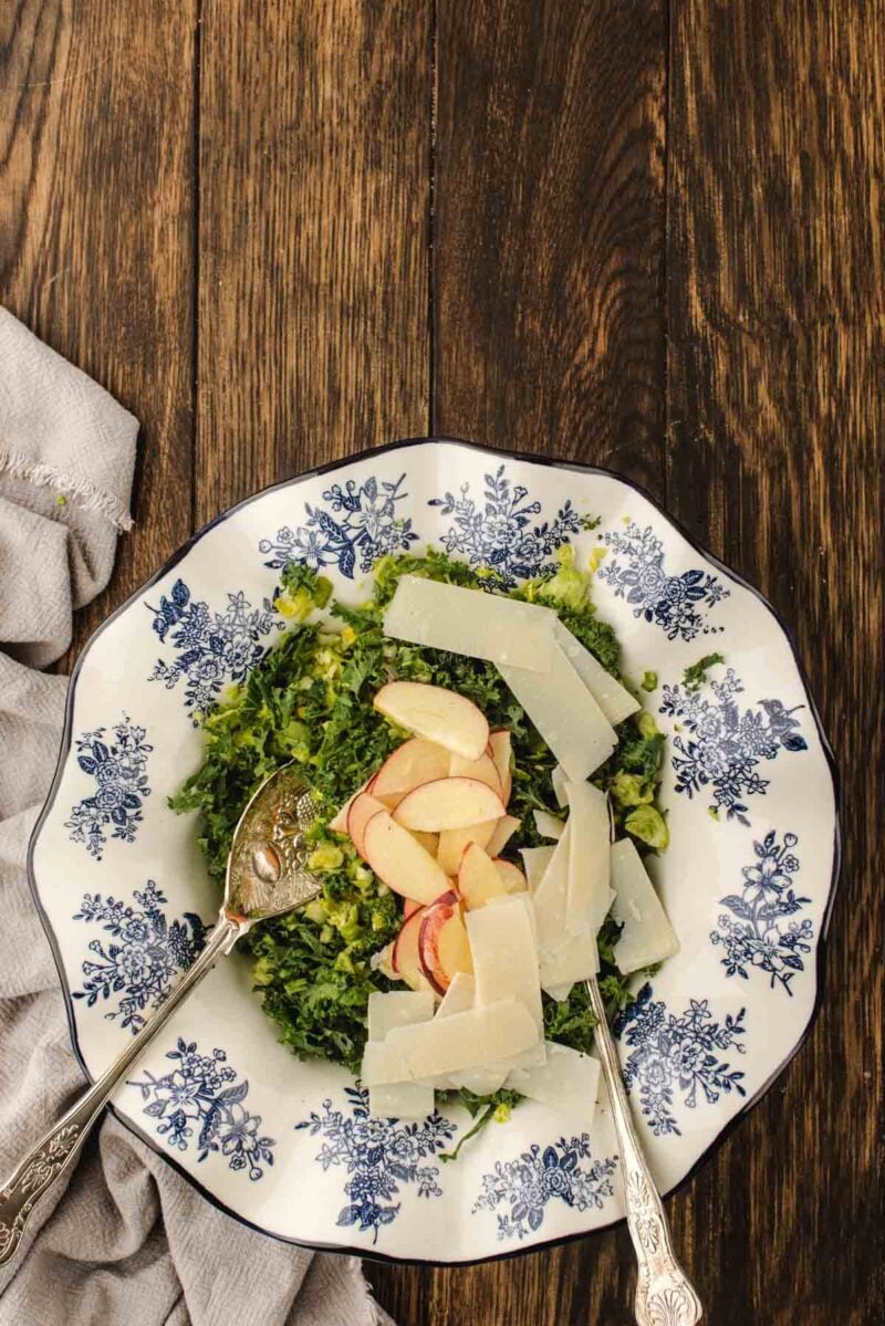 Salad with chopped greens, apple slices, and shaved cheese is served in a white bowl with blue floral patterns on a wooden table, with two serving utensils.