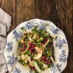 A plate of brussels sprouts kale salad with pomegranate seeds, apple slices, cheese, and pine nuts, served with two spoons on a wooden table.