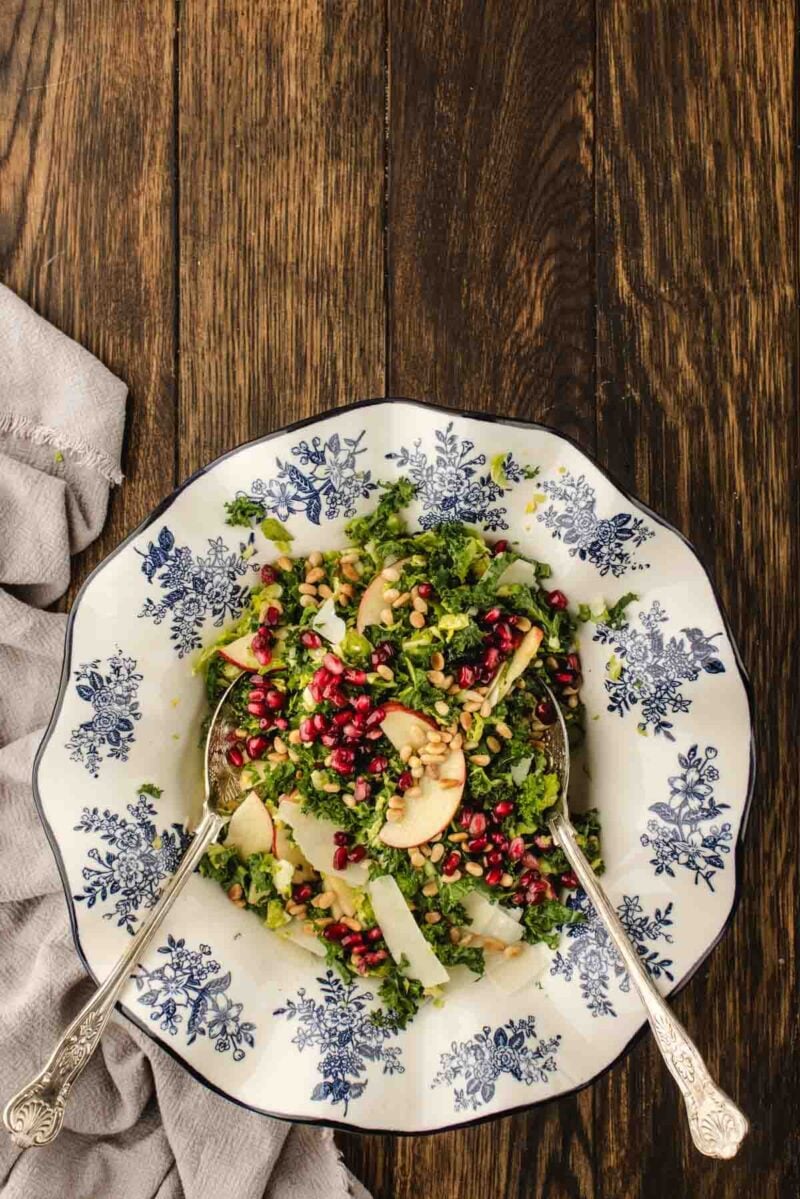 A plate of brussels sprouts kale salad with pomegranate seeds, apple slices, cheese, and pine nuts, served with two spoons on a wooden table.