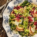 Salad with sliced apples, pomegranate seeds, shaved cheese, and greens on a blue and white patterned plate, with serving utensils beside it.