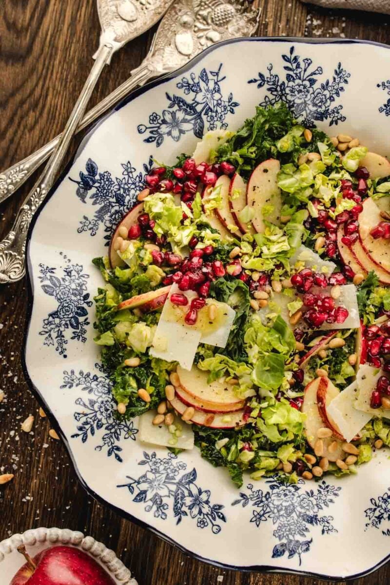 Salad with sliced apples, pomegranate seeds, shaved cheese, and greens on a blue and white patterned plate, with serving utensils beside it.