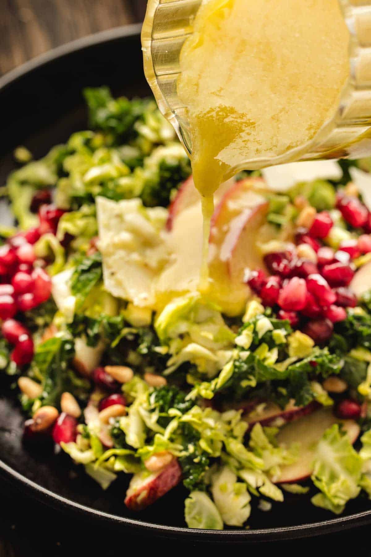 Dressing is being poured over a brussels sprouts salad with leafy greens, apple slices, pomegranate seeds, and pine nuts on a black plate.