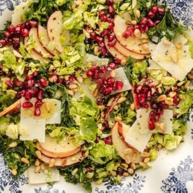 A brussels sprouts salad with sliced apples, pomegranate seeds, shaved cheese, chopped greens, and pine nuts served on a decorative plate.