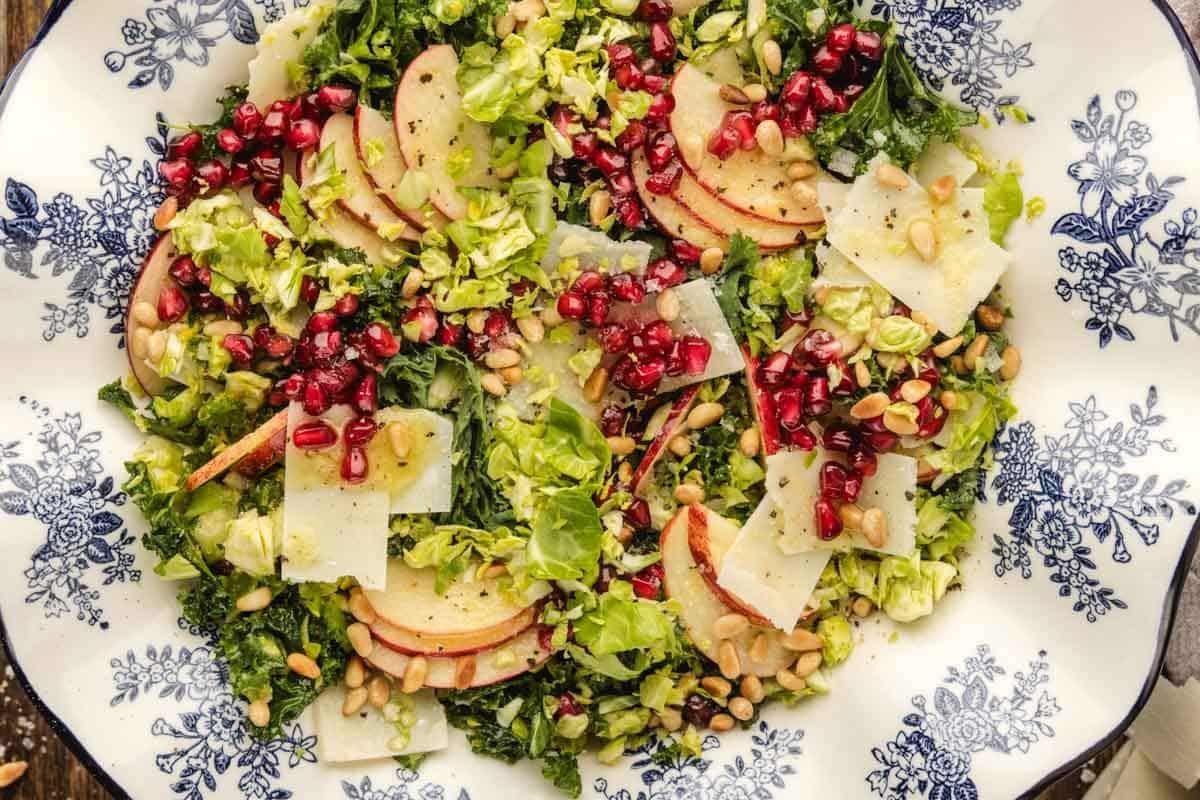 A brussels sprouts salad with sliced apples, pomegranate seeds, shaved cheese, chopped greens, and pine nuts served on a decorative plate.