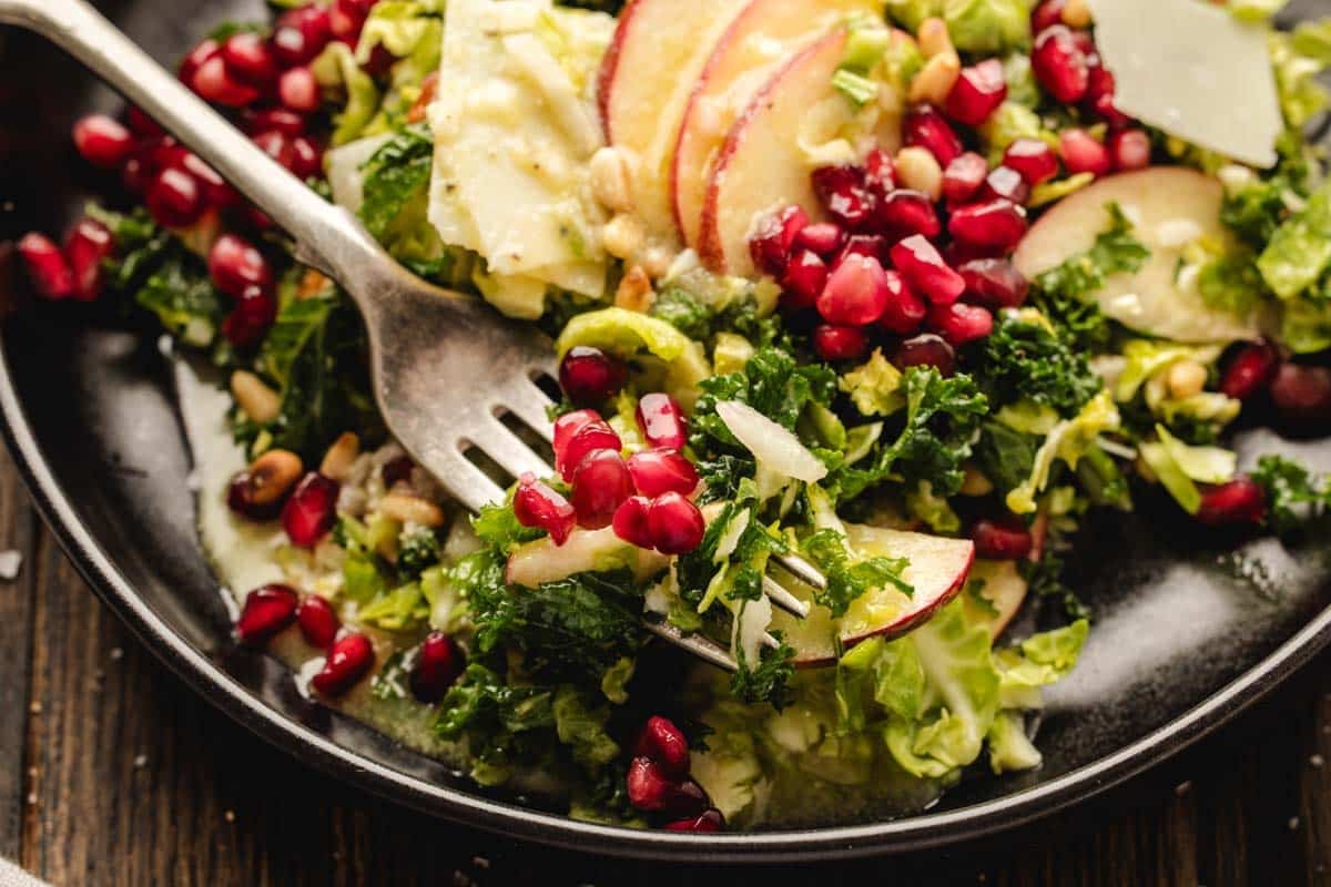 A fork rests on a plate of salad with kale, lettuce, apple slices, pomegranate seeds, and cheese.