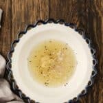 White bowl with scalloped blue rim containing oil, vinegar, chopped shallots, mustard, and pepper on a wooden surface with a beige cloth nearby.