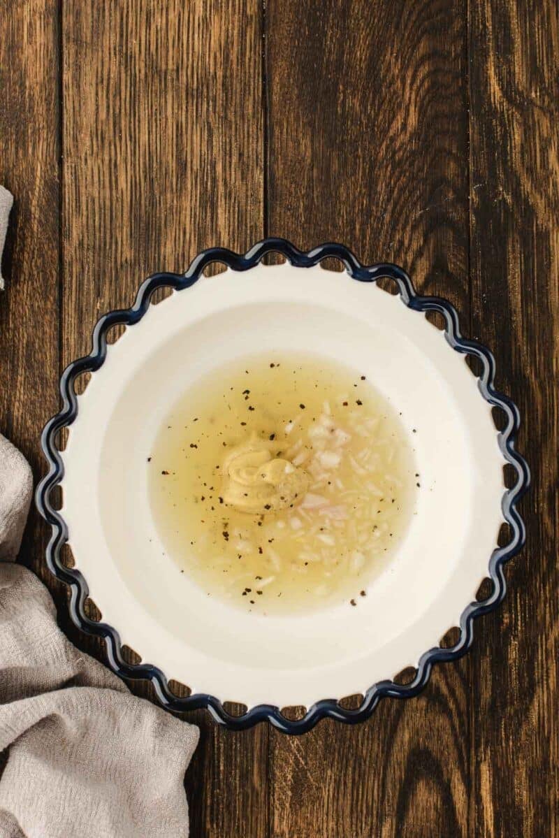 White bowl with scalloped blue rim containing oil, vinegar, chopped shallots, mustard, and pepper on a wooden surface with a beige cloth nearby.