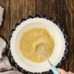 A hand whisks a yellow sauce with visible herbs in a white bowl with a blue scalloped edge, set on a wooden surface next to a beige cloth.