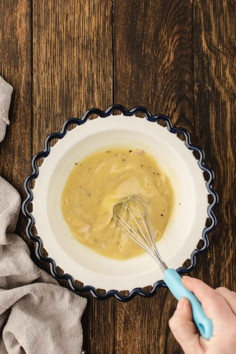 A hand whisks a yellow sauce with visible herbs in a white bowl with a blue scalloped edge, set on a wooden surface next to a beige cloth.