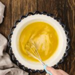 A hand whisking a yellow mixture in a scalloped-edge bowl on a wooden surface, with a beige cloth nearby.