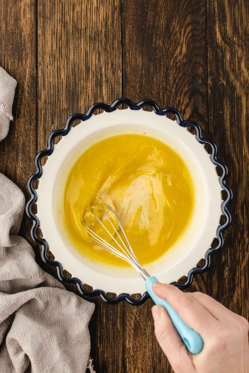 A hand whisking a yellow mixture in a scalloped-edge bowl on a wooden surface, with a beige cloth nearby.