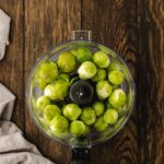 A food processor bowl filled with raw brussels sprouts sits on a wooden table next to a beige cloth.