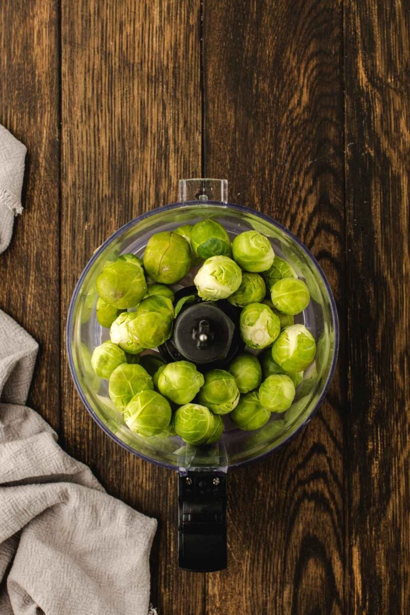 A food processor bowl filled with raw brussels sprouts sits on a wooden table next to a beige cloth.