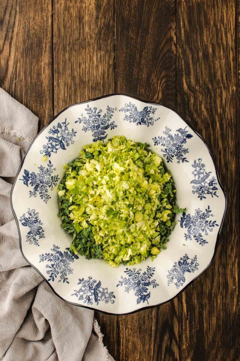 Chopped Brussels sprouts in a white bowl with blue floral designs, placed on a wooden table next to a light-colored cloth.