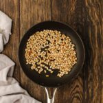A black frying pan with toasted pine nuts sits on a dark wooden surface next to a light gray cloth.