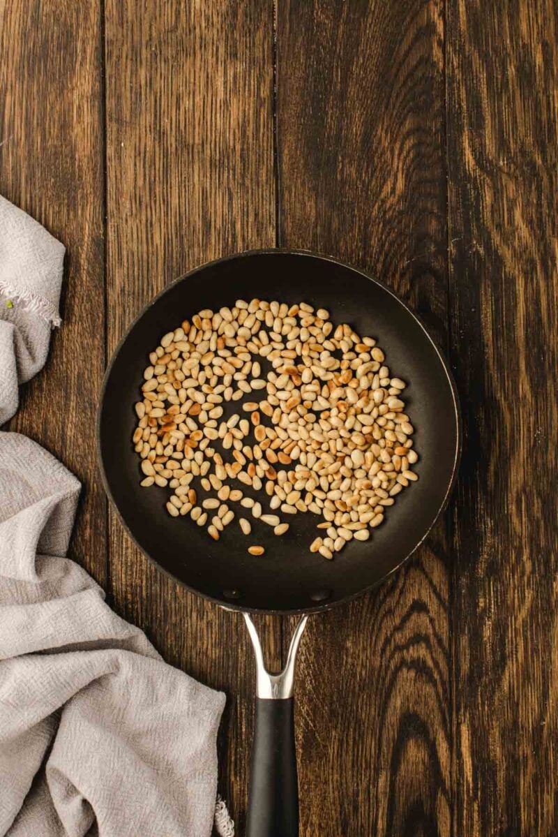 A black frying pan with toasted pine nuts sits on a dark wooden surface next to a light gray cloth.