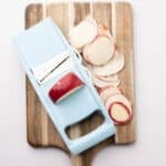 A light blue mandoline slicer and thinly sliced apples on a wooden cutting board against a white background.