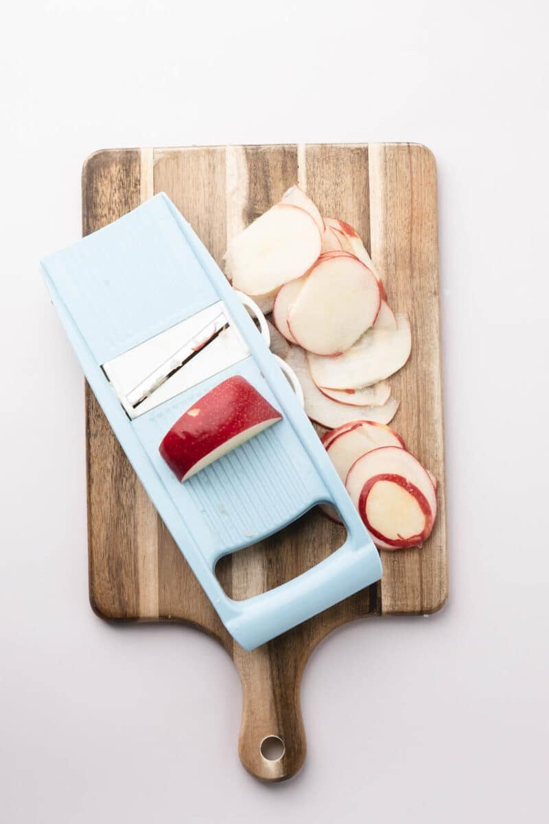 A light blue mandoline slicer and thinly sliced apples on a wooden cutting board against a white background.