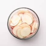A glass bowl filled with thinly sliced red apples on a white background.