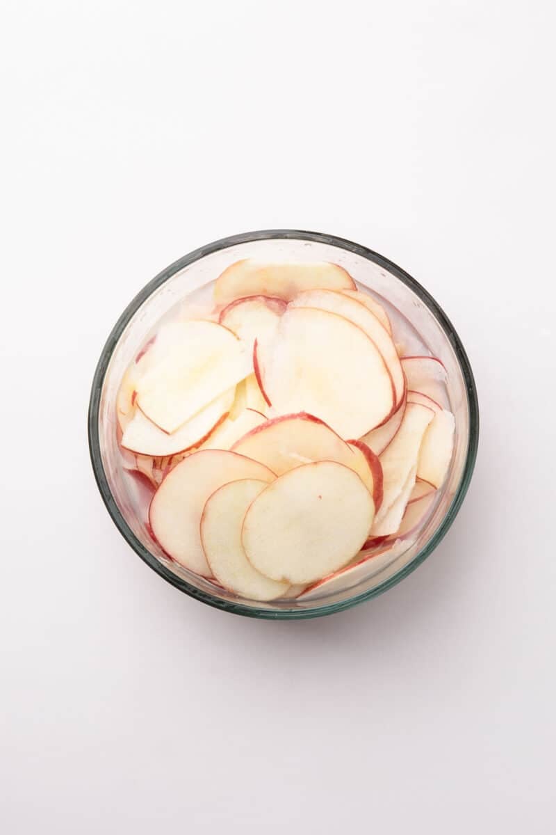 A glass bowl filled with thinly sliced red apples on a white background.