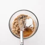 A glass bowl with brown sugar, cornstarch, cinnamon, and liquid ingredients being mixed with a metal whisk on a white background.