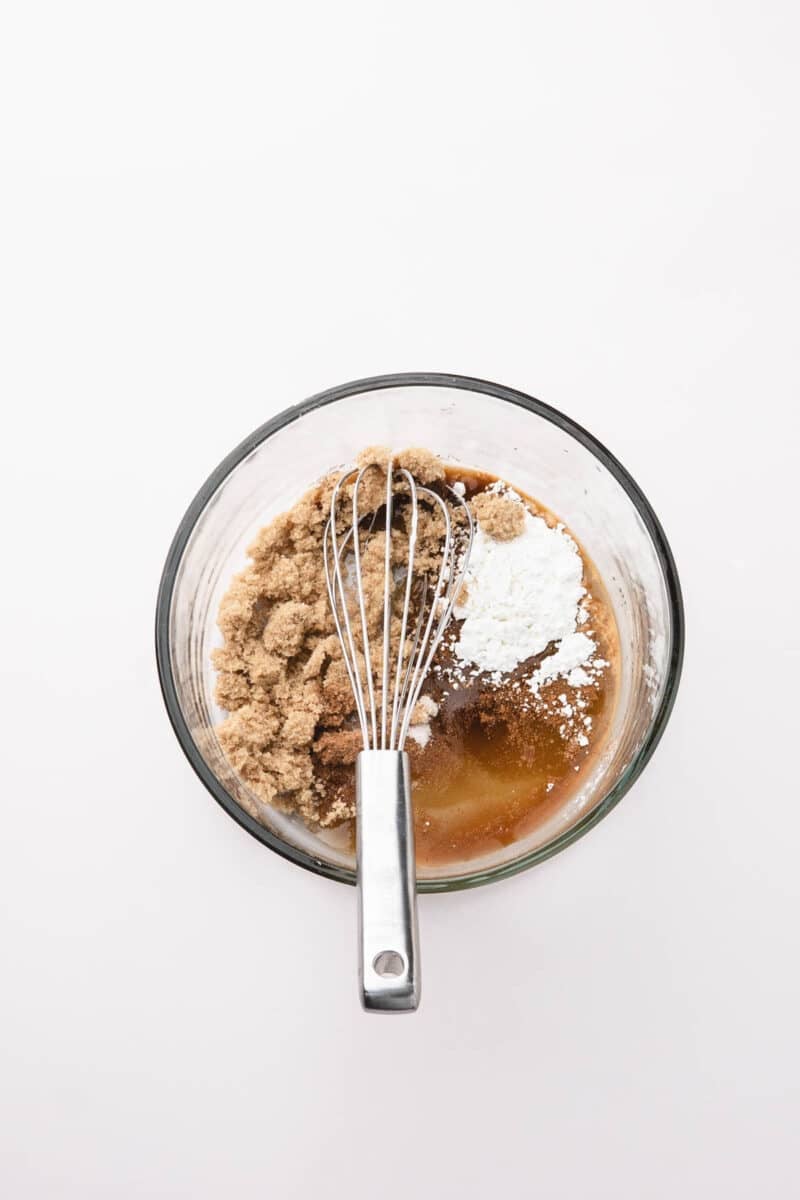 A glass bowl with brown sugar, cornstarch, cinnamon, and liquid ingredients being mixed with a metal whisk on a white background.