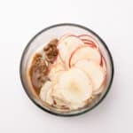 A glass bowl containing thinly sliced apples and a portion of brown sugar mixture on a white background.