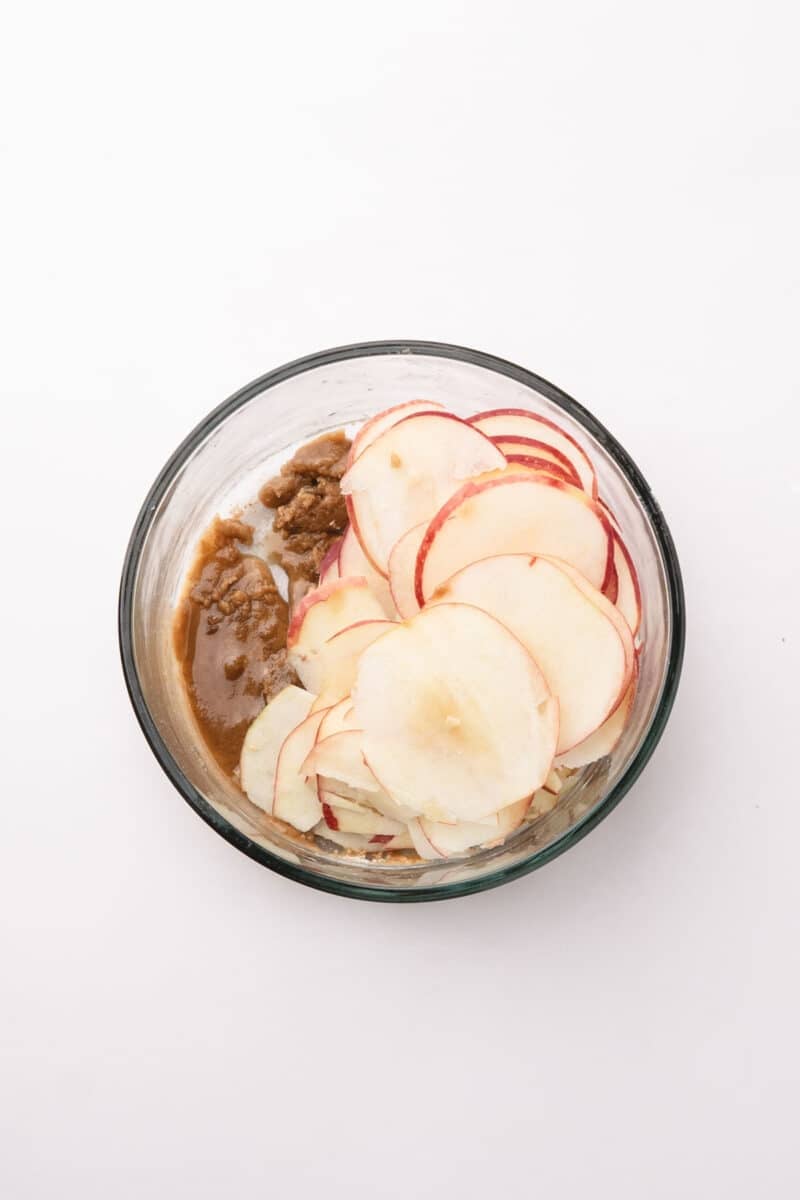 A glass bowl containing thinly sliced apples and a portion of brown sugar mixture on a white background.