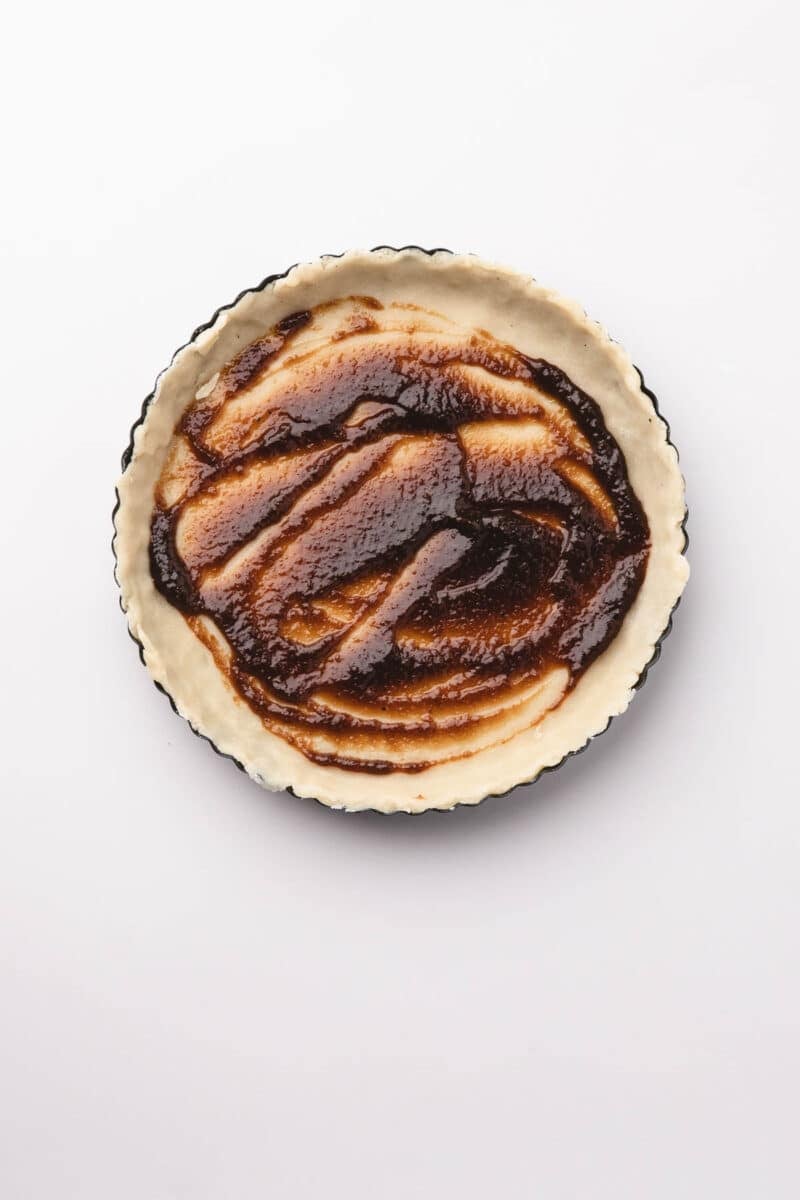 Unbaked pie crust in a round pan, spread with an even layer of brown filling or sauce on top, set against a white background.