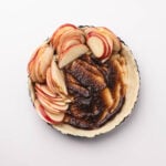 A pie crust filled with brown sugar filling, topped with thinly sliced red apples arranged on one side, on a white background.