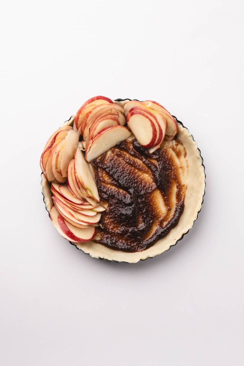 A pie crust filled with brown sugar filling, topped with thinly sliced red apples arranged on one side, on a white background.