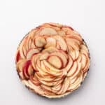 A tart pan filled with thinly sliced red-skinned apples arranged in overlapping concentric circles on a white background.