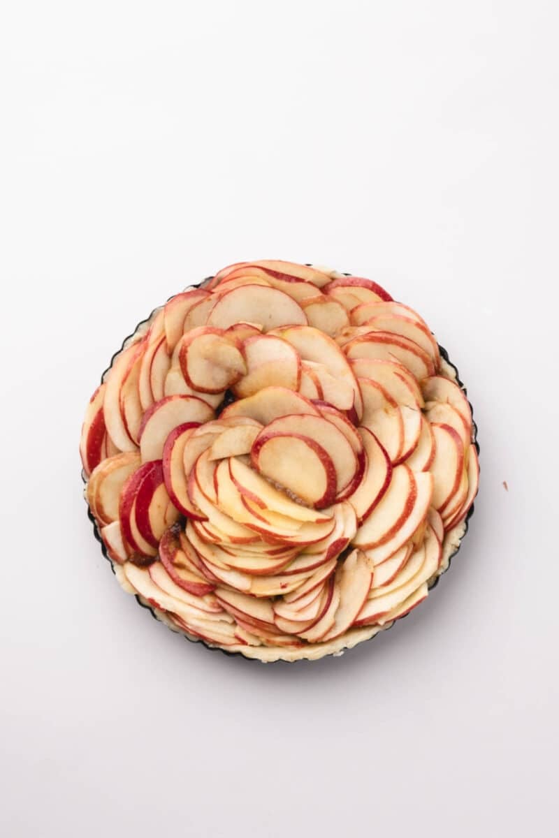A tart pan filled with thinly sliced red-skinned apples arranged in overlapping concentric circles on a white background.