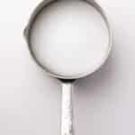 A metal saucepan filled with granulated white sugar is shown from above on a white background.