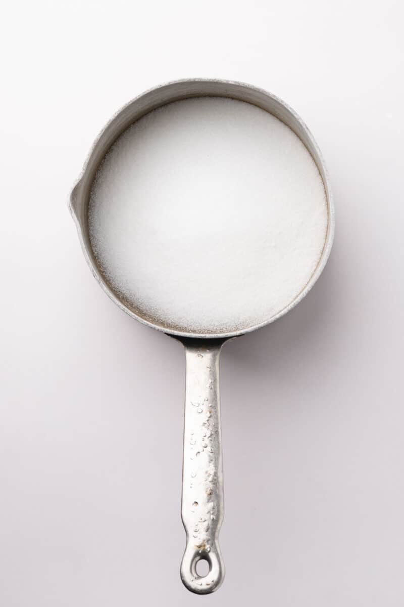 A metal saucepan filled with granulated white sugar is shown from above on a white background.