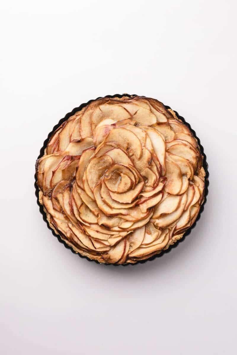 A round tart with thinly sliced apples arranged in a rose pattern, photographed from above on a white background.