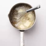 A saucepan with melting butter and a whisk inside, viewed from above, on a plain white background.