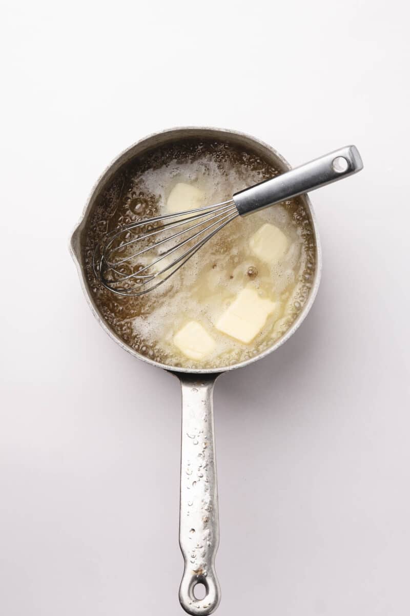 A saucepan with melting butter and a whisk inside, viewed from above, on a plain white background.