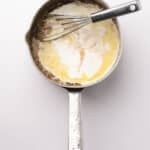 A metal saucepan with melted butter, cream, and a whisk inside, photographed from above on a white background.