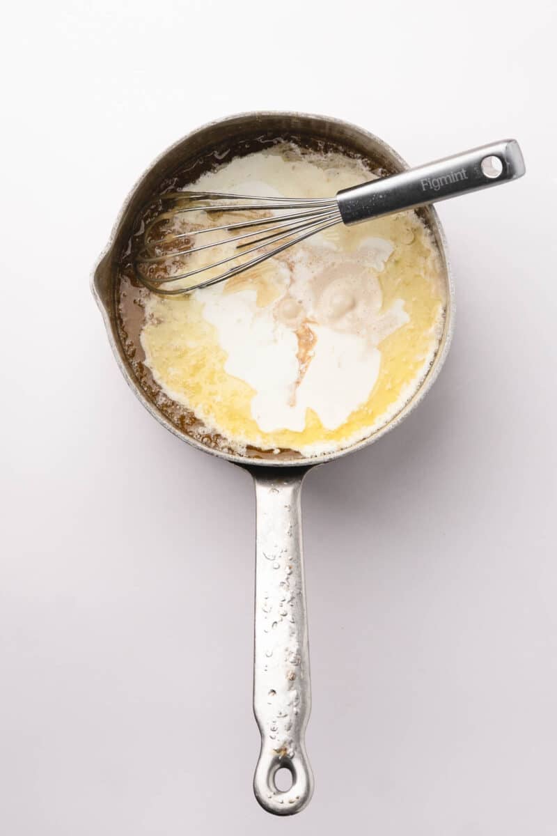 A metal saucepan with melted butter, cream, and a whisk inside, photographed from above on a white background.