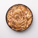 A round caramel apple tart with thinly sliced apples arranged in a spiral pattern and drizzled with caramel glaze, in a black tart pan on a white background.